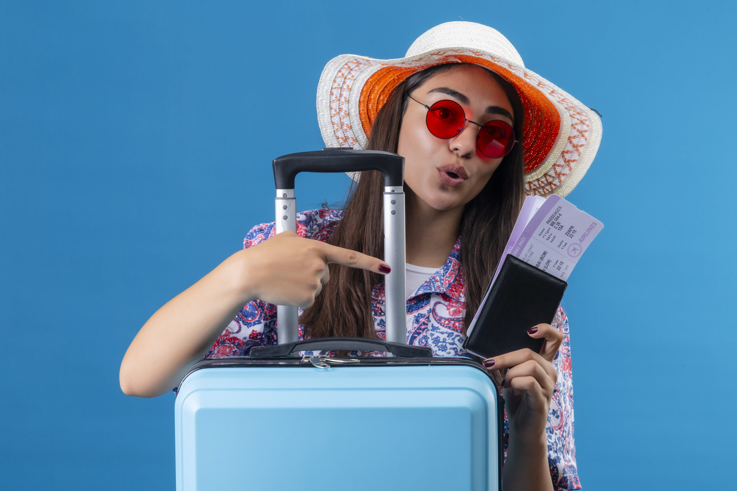 Home young beautiful woman tourist wearing summer hat and red sunglasses holding travel suitcase and tickets with passport pointing with finger to the looking positive and happy standing over blue b